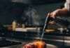 Ein dramatisches Foto einer "Trüffel Beef Roll" , die gerade mit einem Flambierbrenner bearbeitet wird. Leichte Rauchschwaden und die glühende Oberfläche erzeugen eine hochwertige, exklusive Atmosphäre