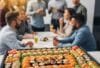 Eine große, rechteckige Sushi-Platte, reich gefüllt mit einer bunten Mischung aus Maki, Nigiri und Special Rolls. Im Hintergrund sind unscharf Menschen in einem Büro-Meeting oder bei einer Feier zu sehen.