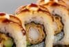 Eine Nahaufnahme einer "Crunchy Golden Palace Roll" , die angeschnitten ist, sodass man die Füllung sieht. Die goldbraune, knusprige Panade ist im Fokus, daneben ein Klecks Teriyakisauce.
